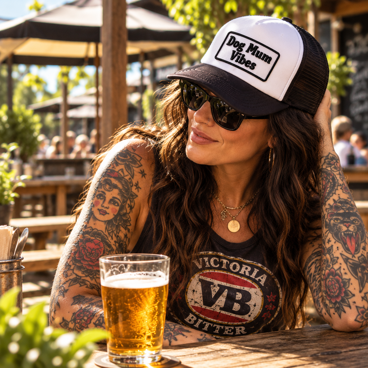 Woman wearing black and white personalised trucker cap with Dog Mum Vibes written on it, sitting in beer garden in a Victoria Bitter singlet with tattoos