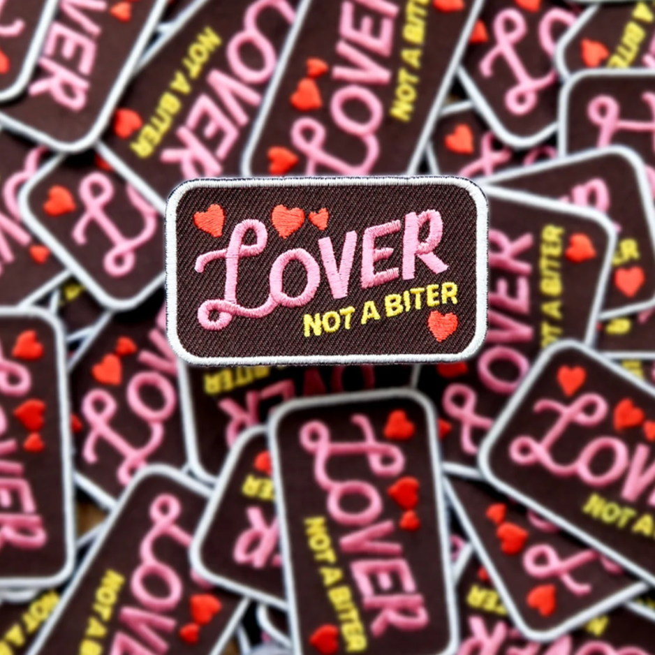 A patch with the text 'Lover not a biter' surrounded by heart shapes in pink color, placed on a background with multiple patches.